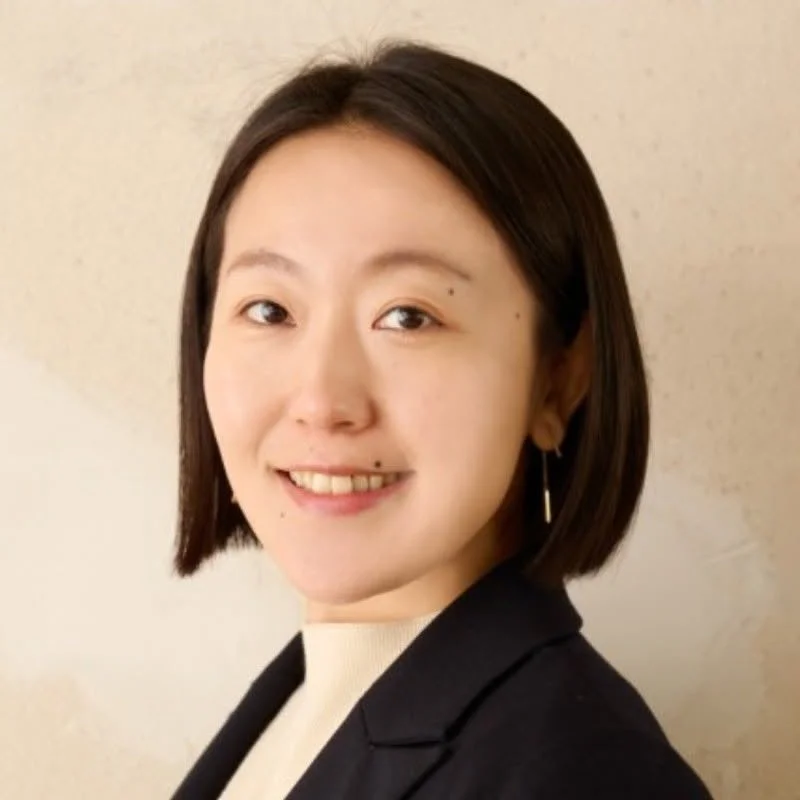 ショートヘアのアジア人女性が、白い壁を背景に穏やかな笑顔を見せている上半身のポートレートです。黒いジャケットと白いインナーを着用しており、プロフェッショナルな印象を与えます。