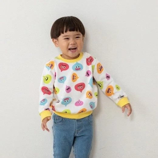 白い壁を背景に、カラフルなキャラクターが描かれたトレーナーとジーンズを着用した小さな男の子が、満面の笑みで立っています。