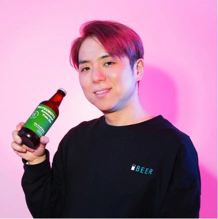 赤い髪の笑顔の男性が、ピンクの背景の前で「MATSUMOTO Awesome Pale Ale」と書かれたビール瓶を持っています。黒い長袖シャツには「BEER」のロゴがあります。