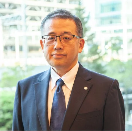 青いスーツと眼鏡をかけたアジア系の中年男性がカメラに向かって立つポートレート。背景は明るいオフィスのようなぼやけた建物で、真面目でプロフェッショナルな雰囲気を醸し出している。