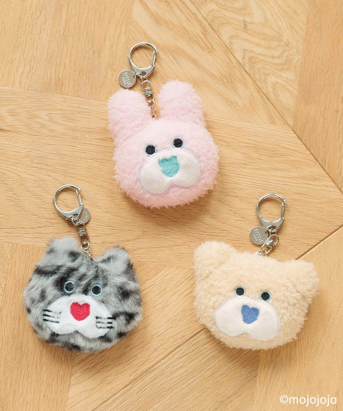 木目の背景に、ピンクのうさぎ、グレーの猫、ベージュのくまのふわふわした動物のキーホルダーが3つ並べられています。可愛らしいデザインが特徴です。