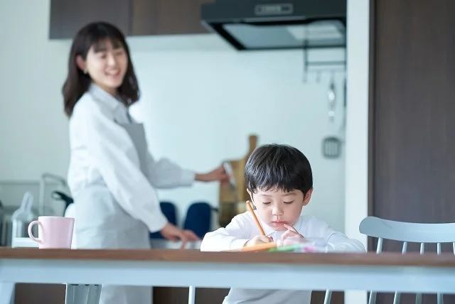 キッチンで家事をする母親のそばで、子供がテーブルで熱心に絵を描いている