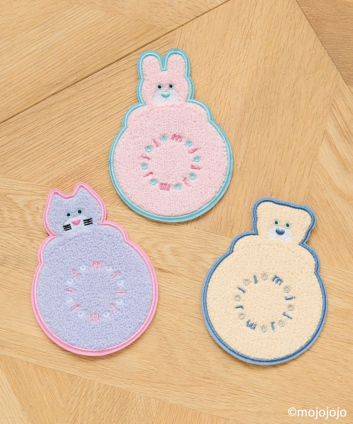 木目のテーブルに、ウサギ、ネコ、クマの形をしたパステルカラーの可愛い刺繍入りコースターが3つ並べられています。それぞれのコースターには「mojomojo」という文字が円形にデザインされており、柔らかな雰囲気を醸し出しています。