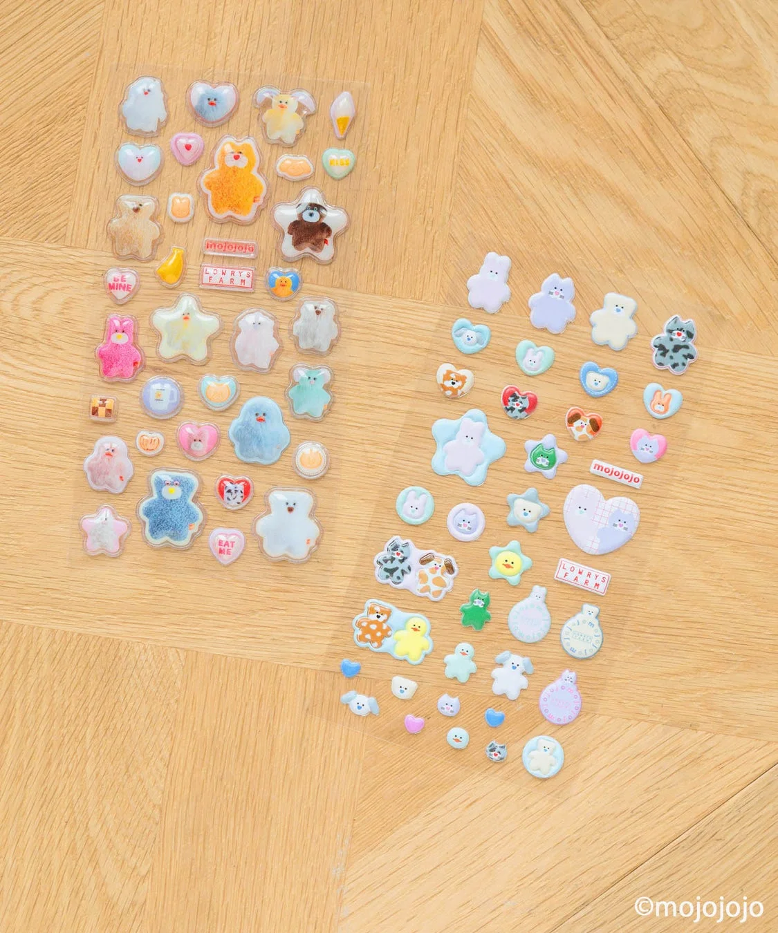 木製の床に、クマやウサギ、ハート、星などの可愛らしいぷっくりとしたステッカーが2シート並べられています。パステルカラーが中心で、全体的に柔らかくキュートな雰囲気です。「mojojojo」や「LOWRYS FARM」などの文字も確認でき、手帳や持ち物のデコレーションにぴったりの雑貨です。