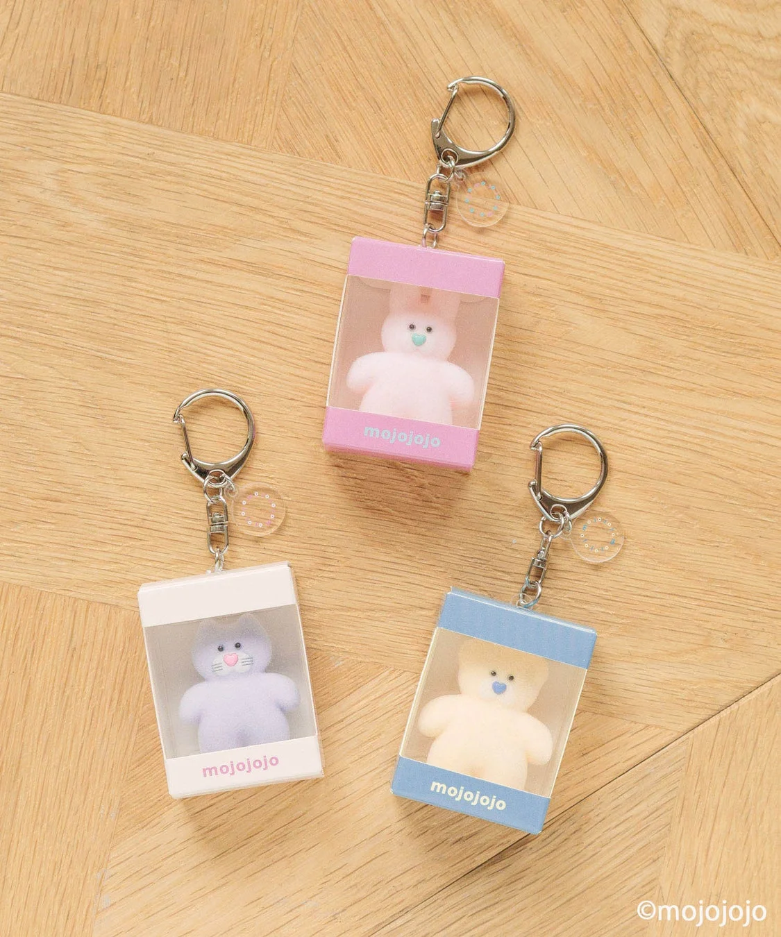 木目の背景に、ピンク、白、水色の箱に入ったうさぎ、猫、くまの可愛いぬいぐるみキーホルダーが3つ並べられています。