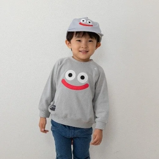 白い背景の前で、笑顔の男の子が、グレーのキャラクターフェイスデザインのトレーナーと帽子を身につけて立っています。