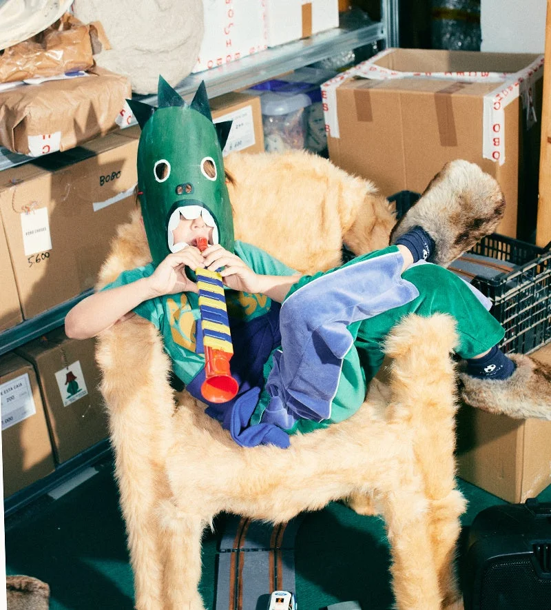緑のモンスターマスクをかぶった子供が、毛皮の椅子に座り、おもちゃの楽器を演奏しています