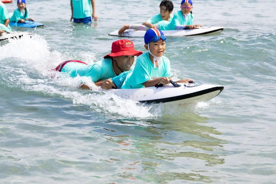 海でインストラクターと一緒にボードに乗って波を楽しむ子供たちの様子