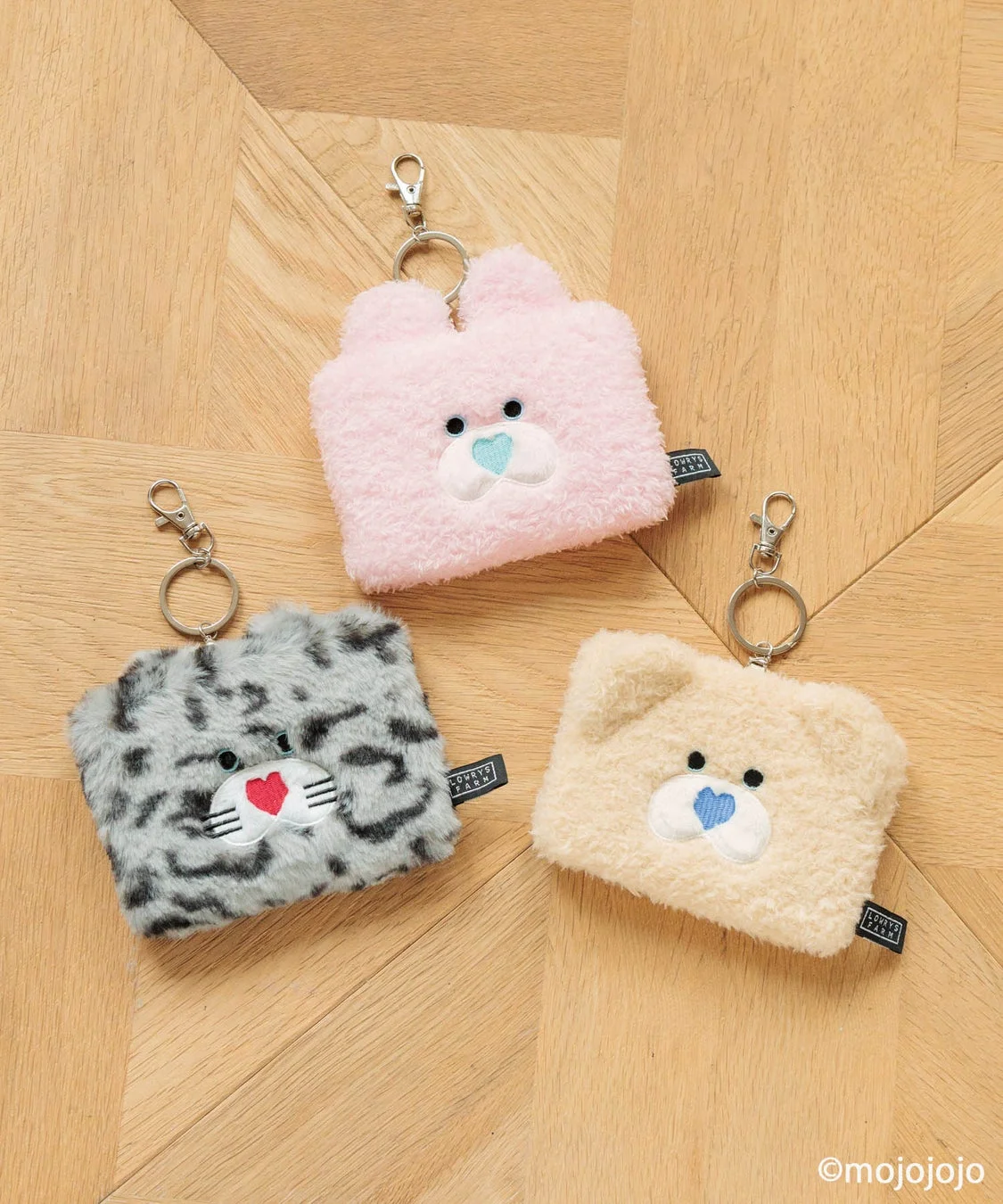 木目の床に、ピンク、ベージュ、グレーの動物モチーフのもこもこしたキーホルダーポーチが3つ並べられています。ウサギ、クマ、ネコ（ヒョウ柄）を模した可愛らしいデザインです。