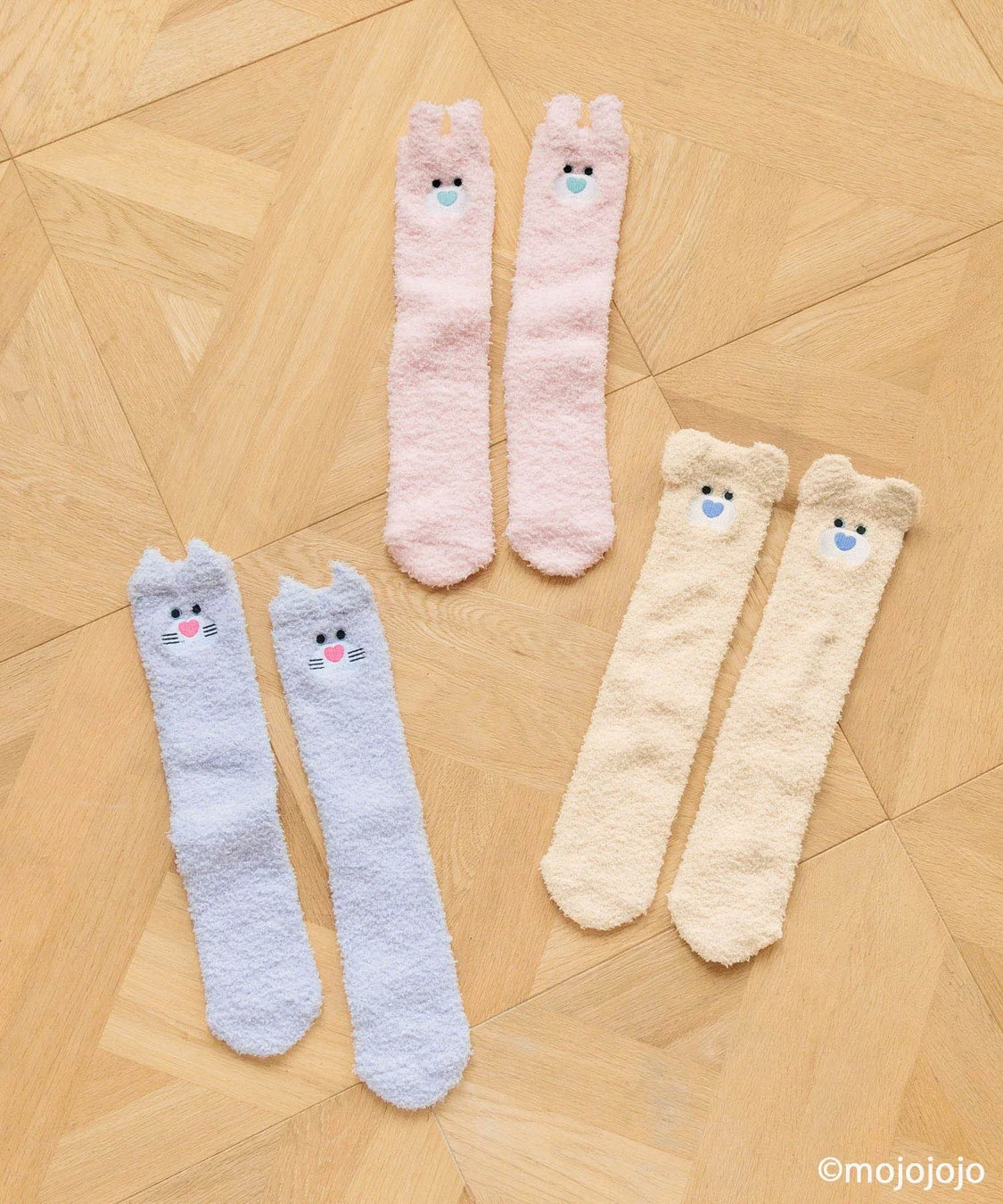 木目の床に置かれた、ピンク、ライトブルー、ベージュの3組のもこもこソックス。それぞれ猫、うさぎ、クマの顔がデザインされており、暖かそうな冬のルームウェアを思わせる可愛らしいアイテムです。