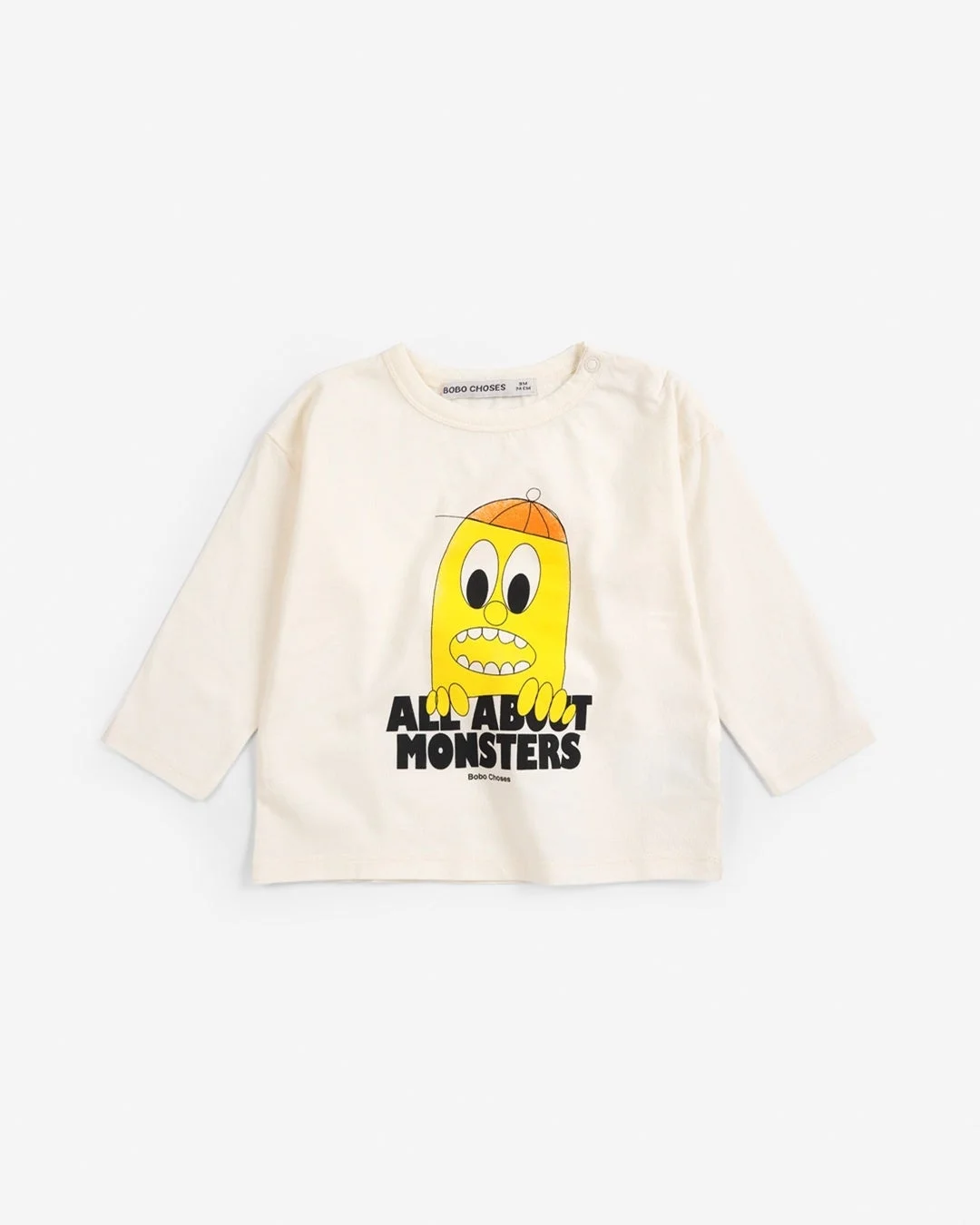 クリーム色の長袖Tシャツで、オレンジ色のキャップをかぶった黄色いモンスターのイラストと「ALL ABOUT MONSTERS」の文字がプリントされています