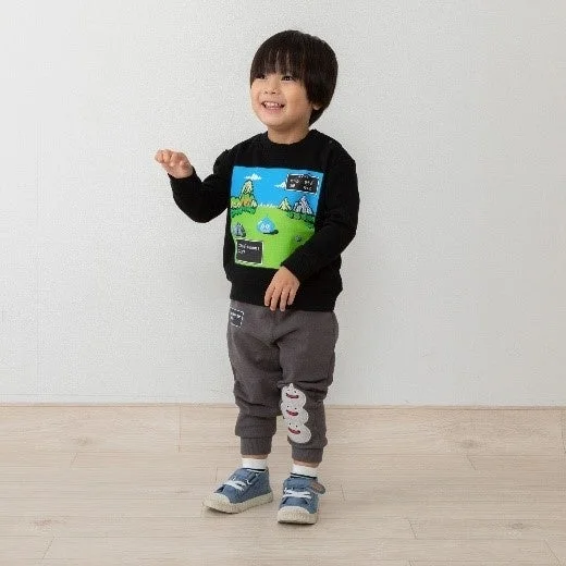 笑顔の男の子が白い壁とフローリングの部屋に立っています。黒いスライム柄のスウェットシャツとグレーのパンツ、青いスニーカーを着用し、左手を少し上げています。