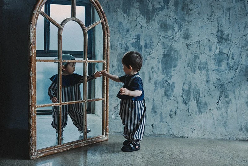ストライプの服を着た幼児が、アンティークなアーチ型鏡に映る自分に手を伸ばしている。