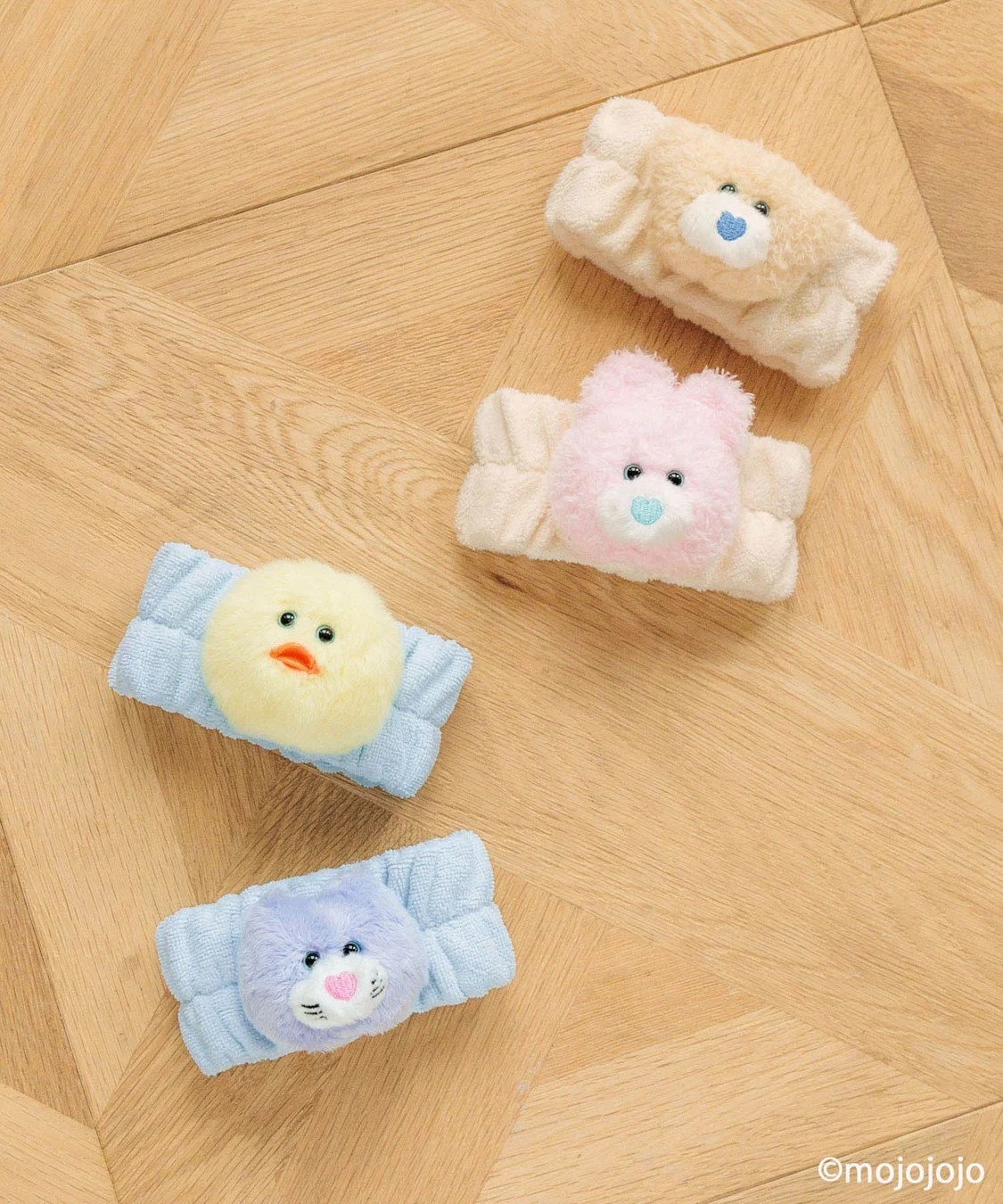 木目の床に、くま、ひよこ、ねこなどのキャラクターがデザインされた、ふわふわで可愛らしい洗顔ヘアバンドが4つ並んでいます。