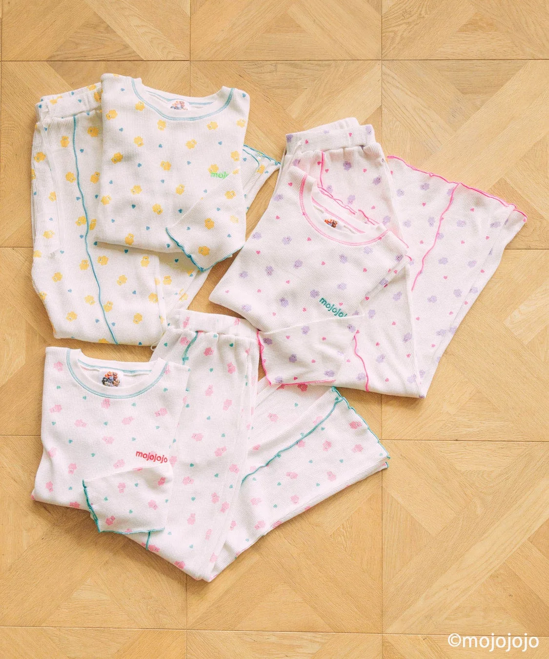 木目の床に、白地にカラフルなドットやハート柄が施された長袖の子供用パジャマが3組、きれいにたたんで置かれている。ベビー・キッズ向けの快適なルームウェアです。