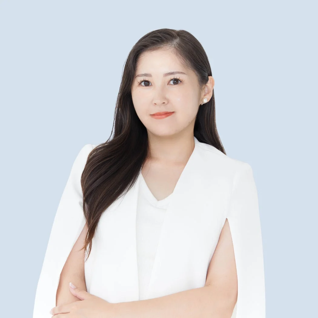 明るい水色の背景に、白いケープ風のジャケットを着用したアジア系の女性のポートレート。長い黒髪で、穏やかな笑顔を浮かべ、腕を組んでいる。清潔感があり、プロフェッショナルな印象を与える。